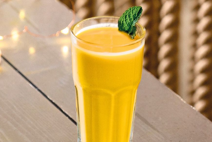 A glass of a yellow liquid, with a leaf of mint sticking out of it